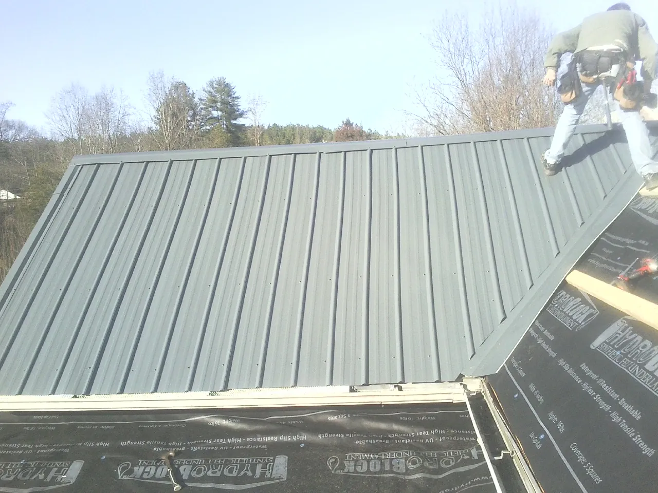 Crew installing dark green standing seam panels over synthetic underlayment in Greenfield